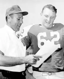 A black and white photo of C. O. Brocato holding a piece of paper and talking to football player Tom Kelly.