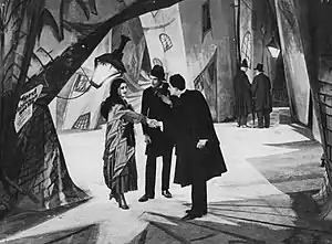 A black-and-white film still.  Three figures stand facing each other on a heavily stylized street set.