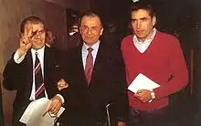 Three men are walking side-by-side holding papers. The first two are wearing a suit and the third is wearing a red sweater. The first man is smiling and flashing a V sign.