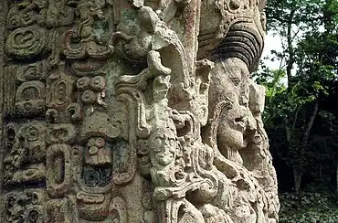 Intricately carved portrait of a human face looking to the right, seen almost in profile against a background of trees. The face is surrounded by highly ornate interlocking designs.