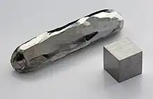 A bar and a cube of a silvery metal crystal lying on a grey surface