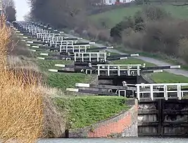 A series of approximately 20 black lock gates with white ends to the paddle arms and wooden railings, each slightly higher than the one below. On the right is a path and on both sides grass and vegetation.