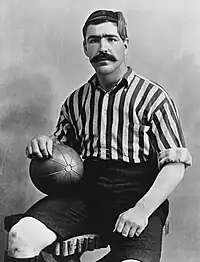 Head and upper torso of a young dark-haired white man with thick eyebrows and a large moustache. He is looking straight ahead, and is wearing a tasselled cap and open-necked striped shirt.