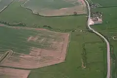 Aerial photograph of fields, with a less-than-straight country lane passing up close to the right hand side. In the top of the frame a modern farmhouse and buildings stand on right of the road. Most of the land is pasture, with two arable fields visible. The crop there is sparse, with large bare patches. The brook wiggles across the upper half of the picture, serpentine in a landscape of straight boundaries. It is narrow and from this height and angle the water surface cannot be discerned. The spring is centre right, its own water course straight and running upwards, toward the brook. The spring is in the greenest of the meadows, with the low humps and bumps of the lost village around.