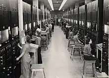 Two rows of control panels with dials and switches. Operators sit at them on four-legged stools.