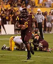 An American football player in a dark red and yellow uniform cradles the football as he sprints towards the camera. Two players in the background lie on the ground.