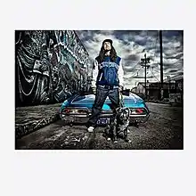 Yankovic in a blue Lynwood jacket standing beside a low-rider convertible with a pit bull on a graffitied urban street scene