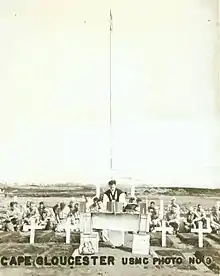 Service personnel with a padre at a memorial service. In the foreground are several graves marked with white crosses.