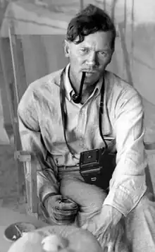 A dark-haired young man, photographed seated, facing the camera and smoking a pipe. His right hand is in a thick glove.