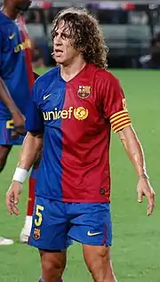 Curly haired man, looking tired, wearing a red and blue football shirt. In the background, green grass is rarely visible.