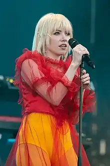 A blonde woman in a red top and yello bottoms holding a microphone