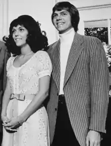 black and white picture of an attractive man and woman in their twenties; the woman on the left is clasping her hands and wearing patterned, short-sleeved dress with a bow, and the man on the right is wearing a striped jacket.