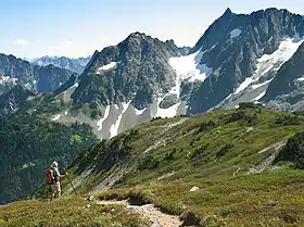 A hiker with walking poles, an orange backpack, and a sun hat, on a trail in a grassed area. In the background are tall dark mountains partly covered in snow.