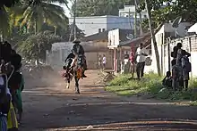 Color photo showing a man on a galloping horse in a dirt street, spectators watching him