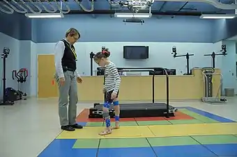 A girl wearing leg braces walks towards a woman in a gym, with a treadmill visible in the background.