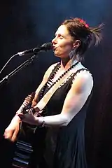 She is shown in upper body shot and left profile. She has her eyes closed as she sings into a microphone and plays her guitar.