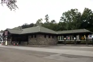 A stone building with a peaked green roof and a porte-cochere on the front. On the left a section of roof extends along some fenced railroad tracks