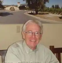 A man with white hair and glasses wearing a brown button-down shirt, smiling at the camera.