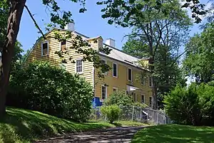 A two-and-a-half-story yellow wooden house with a gambrel roof seen from a corner angle amid large trees. A paved path leading to it from the camera is blocked by a chainlink fence.