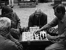 Photo shows two men playing chess while two more look on.