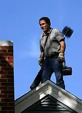 A caucasian man of about forty stands on the ridge of a modern house next to a red brick chimney. He is in jeans and a polo shirt and wears leather safety gloves.On his back is a standard chimney sweeping brush and poles.