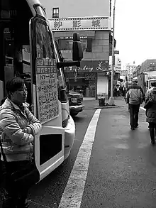 The black-and-white image shows. To the left of the image, a  woman of East Asian extraction standing next to a coach with a sign on its door advertising prices of journeys from New York to Washington D. C. in Han characters. On the right, pedestrians walk across the road in front of the coach under the guidance of a green traffic light. In the left background on the street corner is a Chinese bakery. In the right background there is visible a crowded sidewalk, alongside which three white delivery trucks are parked, and a street of square high-rise apartment buildings.