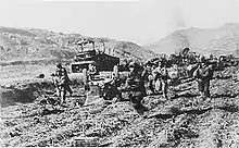 A group of soldier running towards the camera, and the background is littered with wrecked vehicles