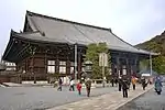 Large wooden building with raised floor, enclosing veranda, white walls and a hip-and-gable roof.