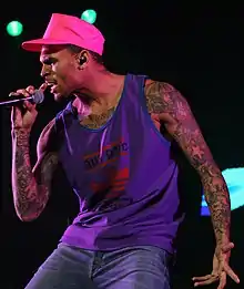 Chris Brown wears a sleeveless purple shirt and a pink hat. He has tattooed arms and he holds a microphone with his right hand, facing to the left.