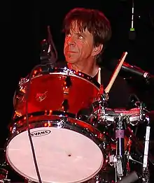 Chris Layton playing a drum kit. He has dark hair and is looking upwards.