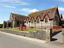 Picture of the  Wesley Methodist church in Front Street, Churchill, taken from the opposite side (south) of the road. The church is shown with red painted window ledges and the cemetery surrounded by red railings.