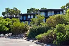 Three dark grey cube-like buildings, each with 2 storeys, are surrounded by native plants.