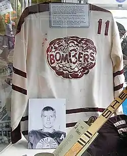 White hockey jersey with maroon trim and a Bombers logo on the chest, accompanied by a hockey stick, a hockey glove, and a photo of Bobby Clarke