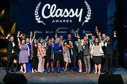 A photograph of twenty people standing in two rows on a stage. They are nicely dressed and holding up award statuettes. The lighting is very blue and behind them is a screen with the Classy logo and sponsors projected.