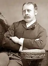 Middle-aged white man with thick moustache, pince-nez glasses and thinning, centre-parted hair, seated, arms folded