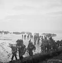 Men in the foreground burdened with equipment, behind them are mud or sand flats and in the distance landing craft