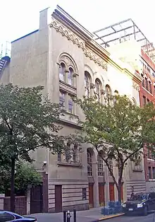 A light brown smooth stone building with two trees in front seen from across and slightly down the street. It has rounded arched windows and decorative moldings at the roofline.