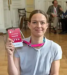 A Caucasian female in a dark blue top, pearl necklace, hair pulled back in a ponytail, holding a microphone.