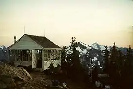 A white-painted wooden building. It has many windows which have wooden covers above them. In the background are mountains.