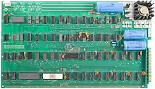 A large, rectangular circuit board with mostly uniform chips arranged neatly in a grid. The rows are labeled A through D and the columns are numbered 1 to 18. Printed between rows of chips is the text, "Apple Computer 1", "Palo Alto, [California] Copyright 1976". There are three large cylindrical capacitors laying sideways in the corner. The board is sprinkled with small components including ceramic resistors and jumper wires.