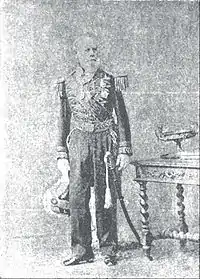 Tintype photograph depicting a full-length portrait of an elderly, bearded man wearing an elaborately embroidered military dress uniform with sword and holding an embroidered and plumed bicorn hat in his right hand