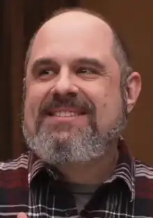 A 51 year-old man with a grey beard smiling to the left of the camera.