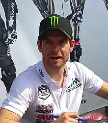 A man sporting a stubble is wearing a black baseball cap with a sponsors logo and a white T-shirt. He is looking away from the camera and holding a blue marker pen in his left hand