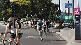 A large number of cyclists ride in the sun near Blackfriars Bridge. There are few cars in sight.