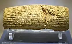 Front view of a barrel-shaped clay cylinder resting on a stand. The cylinder is covered with lines of cuneiform text