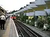 A group of people standing on a railway platform with a railway track to the right and a white-and-red train running on it with its headlights on