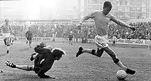 A dramatic action shot from an association football game. A goalkeeper in dark colours forlornly dives to the ground as a tall, lanky forward in a light shirt and white shorts takes the ball around him, controlling it with the outside of his right foot.