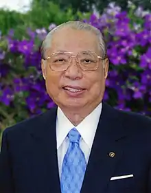 Daisaku Ikeda in a dark jacket, white shirt, light blue tie. Purple flowers fill the background.