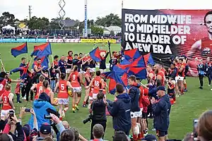 Pearce running towards a banner bearing her portrait followed by her teammates, with Melbourne AFL players and staff forming a guard of honour and fans visible in the background