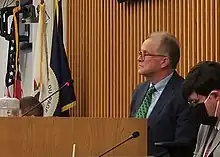 Dan Cronin seated on the dais of the board room. The flags of the United States, the State of Illinois, and County of DuPage are in the background.